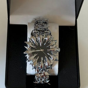 May include: A unique silver-toned wristwatch with a black watch face, presented in a black box. The watch features a spiky, starburst-like design around the face and on the band, creating a bold, avant-garde aesthetic. The watch is displayed on a white cushion.