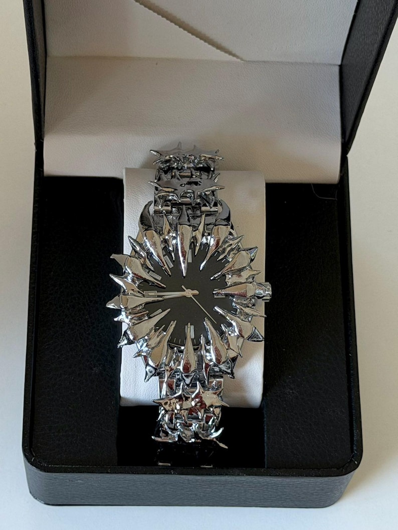 May include: A unique silver-toned wristwatch with a black watch face, presented in a black box. The watch features a spiky, starburst-like design around the face and on the band, creating a bold, avant-garde aesthetic. The watch is displayed on a white cushion.