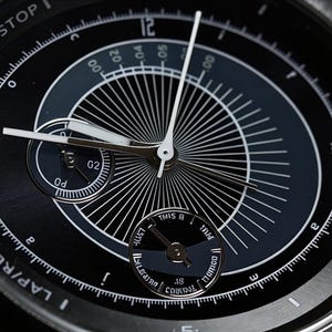 May include: A detailed shot of a black wristwatch with white hands and numerals. The watch face has a complex design with subdials and a radial pattern. The bezel displays the text "START/STOP" and "LAP/RESET".