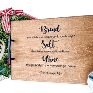 May include: A wooden serving tray with black metal handles. The tray has a quote about bread, salt, and wine, with the text "Bread that this house may never know hunger Salt that life may always have flavor Wine that joy and prosperity may reign forever -W's Wonderful Life-"