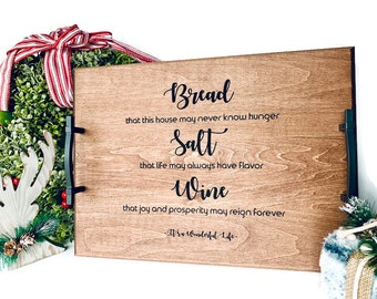 May include: A wooden serving tray with black metal handles. The tray has a quote about bread, salt, and wine, with the text "Bread that this house may never know hunger Salt that life may always have flavor Wine that joy and prosperity may reign forever -W's Wonderful Life-"