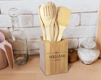May include: A bamboo utensil holder with a set of wooden spoons and spatulas. The holder is engraved with the name "WILLIAMS". The kitchen tools are light brown, and the holder is a natural bamboo color. A clear glass bottle and a pink pitcher are in the background.