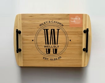 May include: A personalized bamboo serving tray with black handles. The tray features a monogram with the initials "W" and the text "Riley & Cayden, Wellsly, Est. 10.22.25". The tray also has a small sticker that says "Thistle & Sash, Custom Gifts".