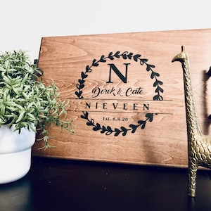 May include: A wooden serving tray with black handles. The tray is personalized with a black wreath design, the letter "N", the names "Derek & Cate", the last name "Nieveen", and the date "Est. 8.8.20". A gold giraffe figurine is on the right side of the tray.