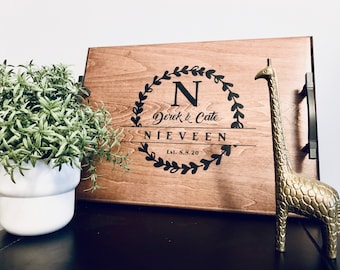 May include: A wooden serving tray with black handles. The tray is personalized with a black wreath design, the letter "N", the names "Derek & Cate", the last name "Nieveen", and the date "Est. 8.8.20". A gold giraffe figurine is on the right side of the tray.