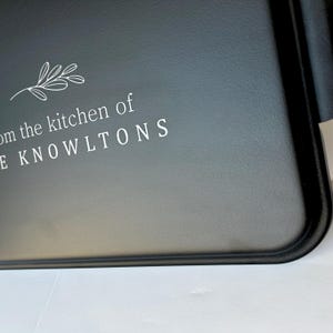 Może przedstawiać: Czarna metalowa taca do serwowania z białym wzorem, na kt&oacute;rym widnieje napis "From the kitchen of the Knowltons" z małym wzorem liścia nad tekstem.