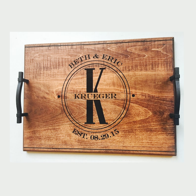 Medium-toned maple wood serving tray. Circle monogram engraved in center, large K in the middle and the name Krueger across. The names Beth and Eric are curved at the top of the circle and the wedding date at the bottom. Text and handles are black.