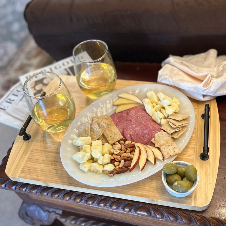 Two-toned bamboo serving tray with charcuterie plate across it, ramekin of olives, and two glasses of white wine.