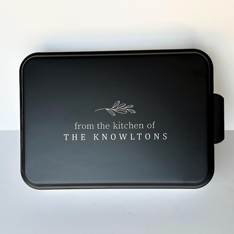 Może przedstawiać: Czarna forma do ciasta z białym wzorem, na kt&oacute;rym widnieje napis "from the kitchen of THE KNOWLTONS" z małym wzorem gałązki nad tekstem.