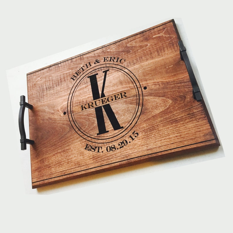 Medium-toned maple wood serving tray. Circle monogram engraved in center, large K in the middle and the name Krueger across. The names Beth and Eric are curved at the top of the circle and the wedding date at the bottom. Text and handles are black.