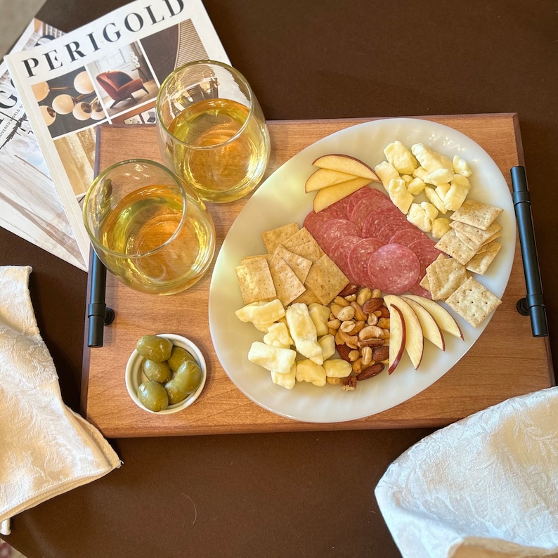 Medium-toned maple wood serving tray with charcuterie plate across it, ramekin of olives, and two glasses of white wine.