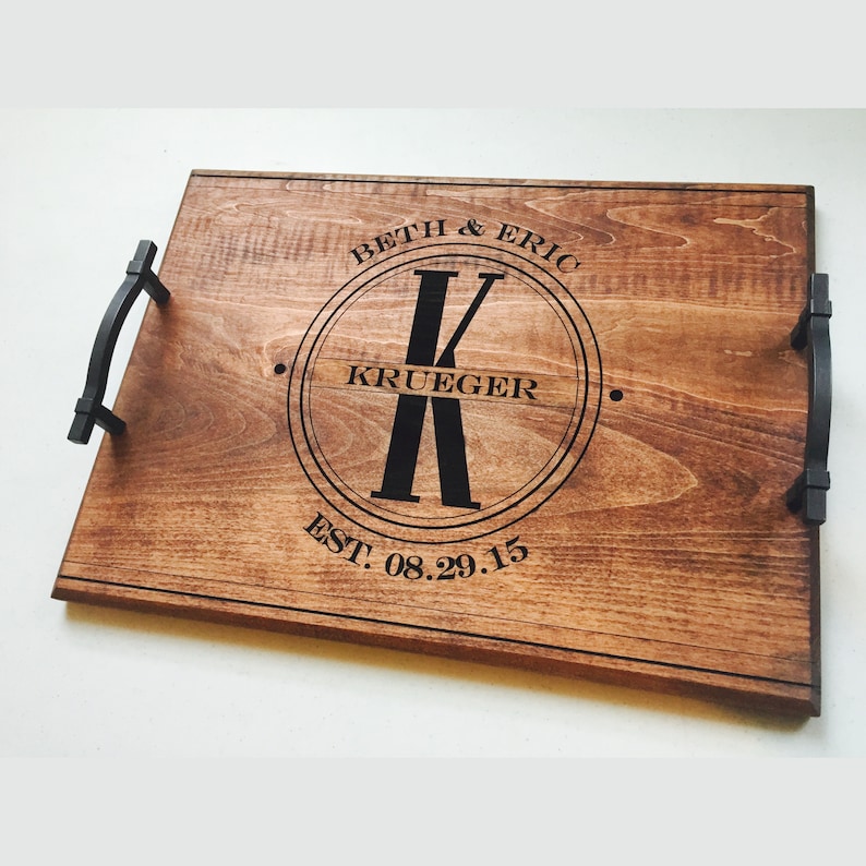 Medium-toned maple wood serving tray. Circle monogram engraved in center, large K in the middle and the name Krueger across. The names Beth and Eric are curved at the top of the circle and the wedding date at the bottom. Text and handles are black.