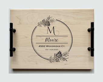 May include: A wooden serving tray with black metal handles. The tray is engraved with a pine branch design, a circle with the letter "M" and the name "Moore", the address "4502 WOODRIDGE CT.", and the date "Est. 4.29.2023".