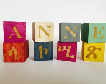 May include: Wooden blocks spelling the name "ANNE" in gold lettering. The blocks are stacked in two rows, with the top row being pink, beige, gray, and green. The bottom row is red, blue, pink, and yellow.
