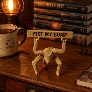 Könnte beinhalten: Eine beige, bewegliche, roboterartige Figur, die auf vier Beinen steht und ihre Arme über den Kopf hebt, um ein rechteckiges Schild zu halten, das den Text "FIST MY BUMP" in dunklen, fetten, serifenlosen Großbuchstaben auf beigem Grund zeigt.