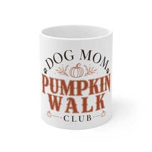 Puede incluir: Una taza de cerámica blanca con el texto en negro y naranja DOG MOM PUMPKIN WALK CLUB. El diseño incluye una ilustración central de una calabaza naranja, pequeños detalles de hojas naranjas y dos huellas de perro negras a los lados.