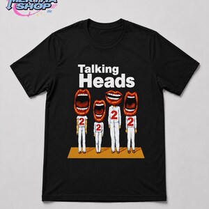May include: A black short-sleeved t-shirt featuring a graphic of four figures with large open mouths instead of heads. Each figure wears a white outfit with a red number 2 on the chest. The text Talking Heads appears in white above the figures.