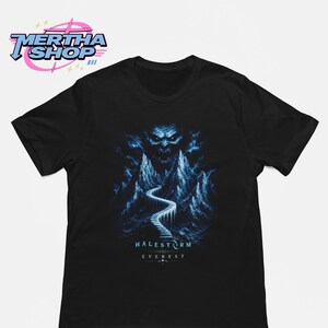 Op de afbeelding: Een zwart t-shirt met korte mouwen en een blauwe afbeelding van een besneeuwd gebergte met een kronkelend pad dat leidt naar een donker, dreigend gezicht dat uit de wolken tevoorschijn komt. De tekst "HALESTORM" en "EVEREST" staat in wit.