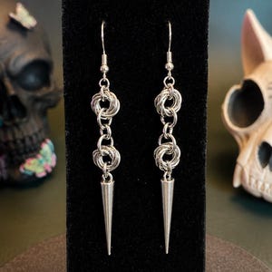 May include: A pair of silver-toned drop earrings featuring a chainmaille design with two interconnected circular links suspended from a hook, terminating in a smooth, pointed metal spike at the base of each earring, displayed on a black stand.