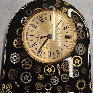 May include: A black clock with a gold-colored face and Roman numerals. The clock is decorated with various gold and silver gears. The clock's hands point to approximately 9:15. The clock has a curved top.