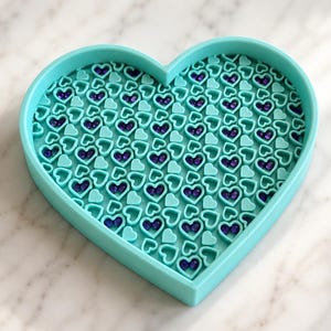 Op de afbeelding: Een hartvormig dienblad in een heldere turquoise kleur. Het binnenoppervlak is gevuld met een herhalend patroon van kleine, verhoogde turquoise hartomtrekken, met piepkleine, donkerpaarse, ronde kraaltjes in veel van de hartvormen.
