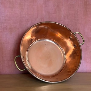 Puede incluir: Un cuenco redondo de cobre pulido con una superficie interior lisa y dos asas curvas de latón fijadas a los lados con remaches visibles. El recipiente tiene una abertura ancha y una base profunda y redondeada de color cobre.