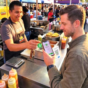 May include: A person holds a smartphone displaying a Cash App payment screen while another person holds a green Cash App key fob near the device. The scene takes place at a food truck with a stainless steel counter, condiment bottles, and tacos.