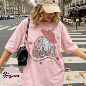 May include: A light pink t-shirt featuring a graphic print of a cowboy boot with a cow print pattern, a red hat, and blocks with the letters T, S, and 13. The design includes the name Taylor, small flowers, stars, and a chain of beads.