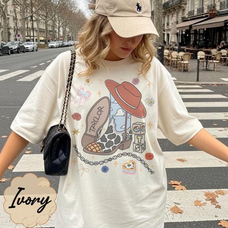 May include: An ivory short-sleeve t-shirt featuring a graphic print of a cowboy boot with a cow print pattern, a red hat, the name Taylor, the number 13, and stacked blocks with the letters T, S, and a heart. A black quilted shoulder bag is worn.