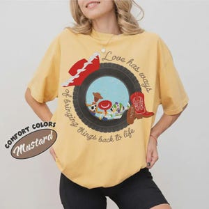 Puede incluir: Una camiseta de manga corta color amarillo mostaza con un gr&aacute;fico de un neum&aacute;tico negro que enmarca personajes de dibujos animados, un sombrero de vaquero rojo y una bota roja. El texto dice Love has ways of bringing things back to life.