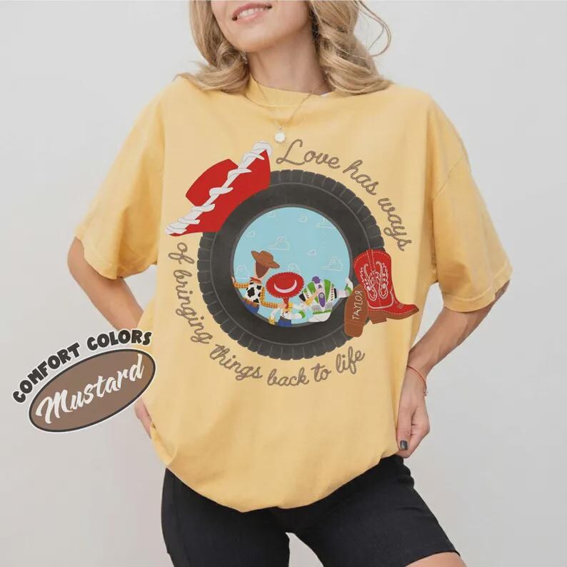 Puede incluir: Una camiseta de manga corta color amarillo mostaza con un gr&aacute;fico de un neum&aacute;tico negro que enmarca personajes de dibujos animados, un sombrero de vaquero rojo y una bota roja. El texto dice Love has ways of bringing things back to life.