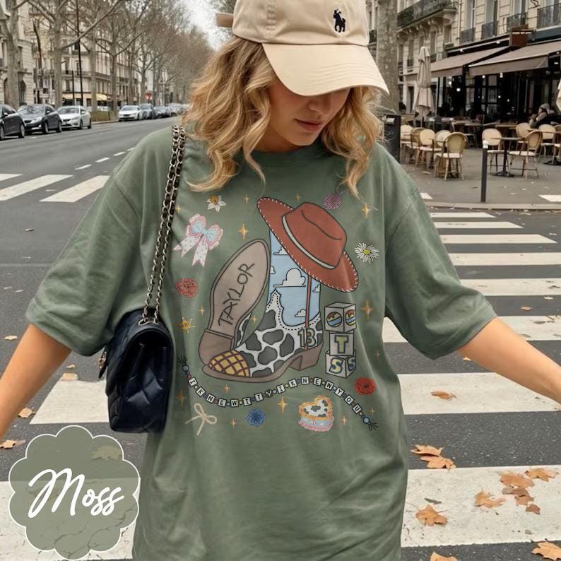 May include: An olive green t-shirt featuring a graphic print of a cowboy boot with a cow print pattern, a brown hat, the name Taylor, the number 13, and various small icons including bows, flowers, and blocks. A black quilted shoulder bag is also visible.