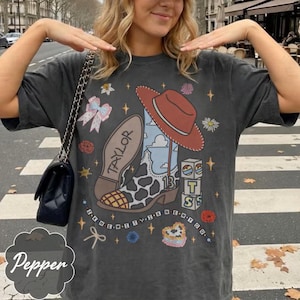 May include: A charcoal gray short-sleeve t-shirt featuring a graphic print of a cowboy boot with a cow print pattern, a red hat, the name Taylor, the number 13, stacked blocks, a bow, flowers, stars, and the text I KNEW IT I KNEW YOU.