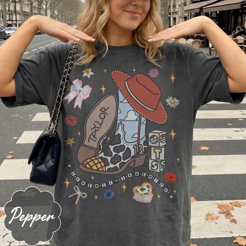 May include: A charcoal gray short-sleeve t-shirt featuring a graphic print of a cowboy boot with a cow print pattern, a red hat, the name Taylor, the number 13, stacked blocks, a bow, flowers, stars, and the text I KNEW IT I KNEW YOU.
