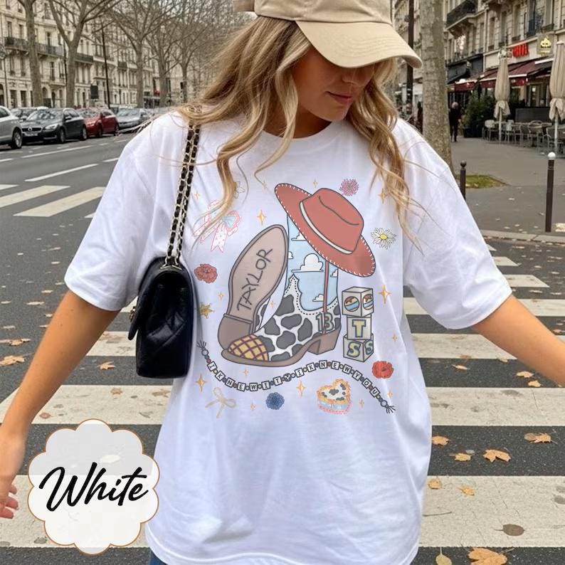 May include: A white t-shirt featuring a graphic print of a cowboy boot with a cow print pattern, a red cowboy hat, and blocks with the letters T, S, and 13. The boot has the name Taylor written on the sole, surrounded by small flowers and stars.
