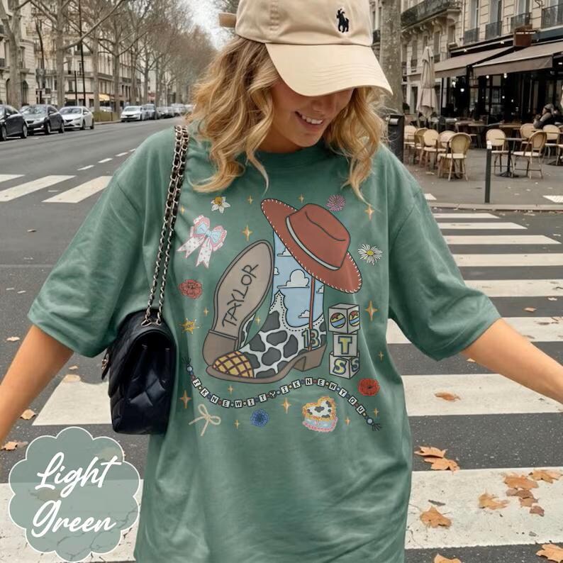 May include: A light green t-shirt featuring a graphic print of a cowboy boot with cow print, a brown hat, a bow, and blocks with the letters T, S, and the number 13. The text Taylor is on the boot sole, and the shirt is paired with a black shoulder bag.