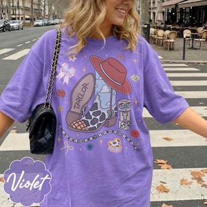 May include: A violet t-shirt featuring a graphic print of a cowboy boot with a cow print pattern, a red hat, and the name Taylor. The design includes blocks with the letters T, S, and the number 13, surrounded by small flowers, bows, and stars.