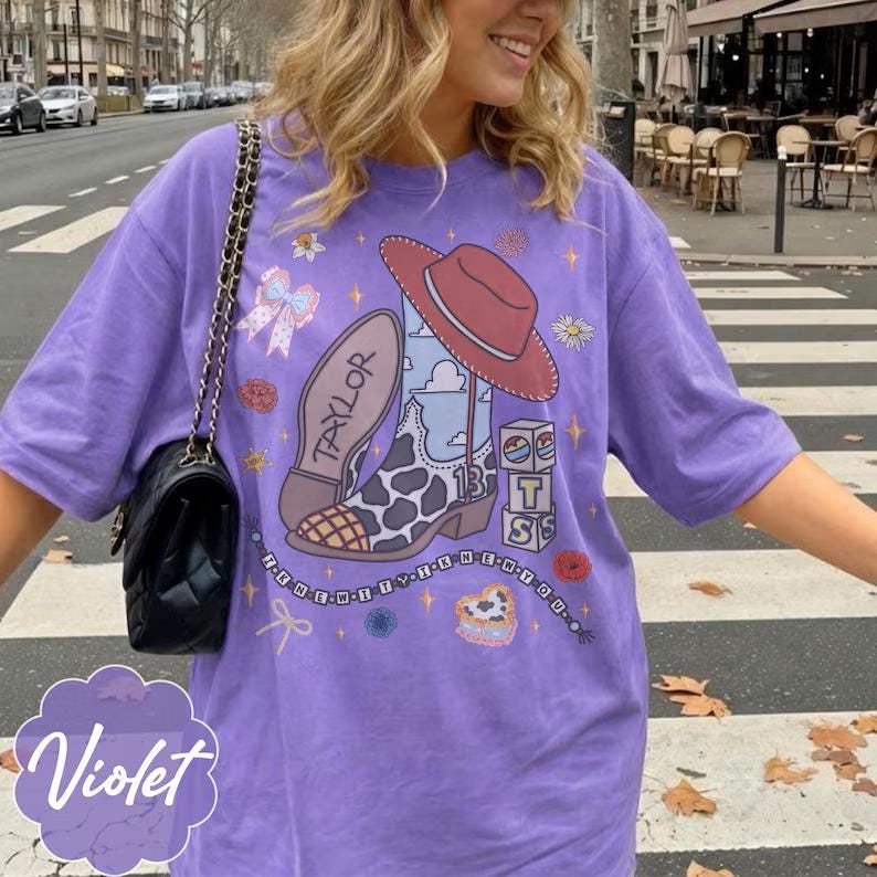 May include: A violet t-shirt featuring a graphic print of a cowboy boot with a cow print pattern, a red hat, and the name Taylor. The design includes blocks with the letters T, S, and the number 13, surrounded by small flowers, bows, and stars.