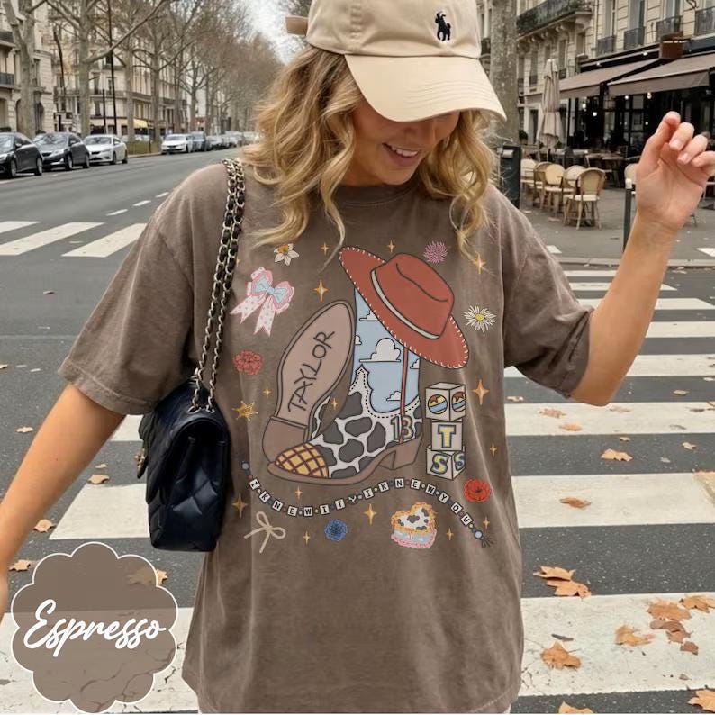 May include: A brown short-sleeve t-shirt featuring a graphic print of a cowboy boot with a cow print pattern, a red hat, a blue sky background, the name Taylor, the number 13, and stacked blocks with the letters T, S, and S, surrounded by small stars.