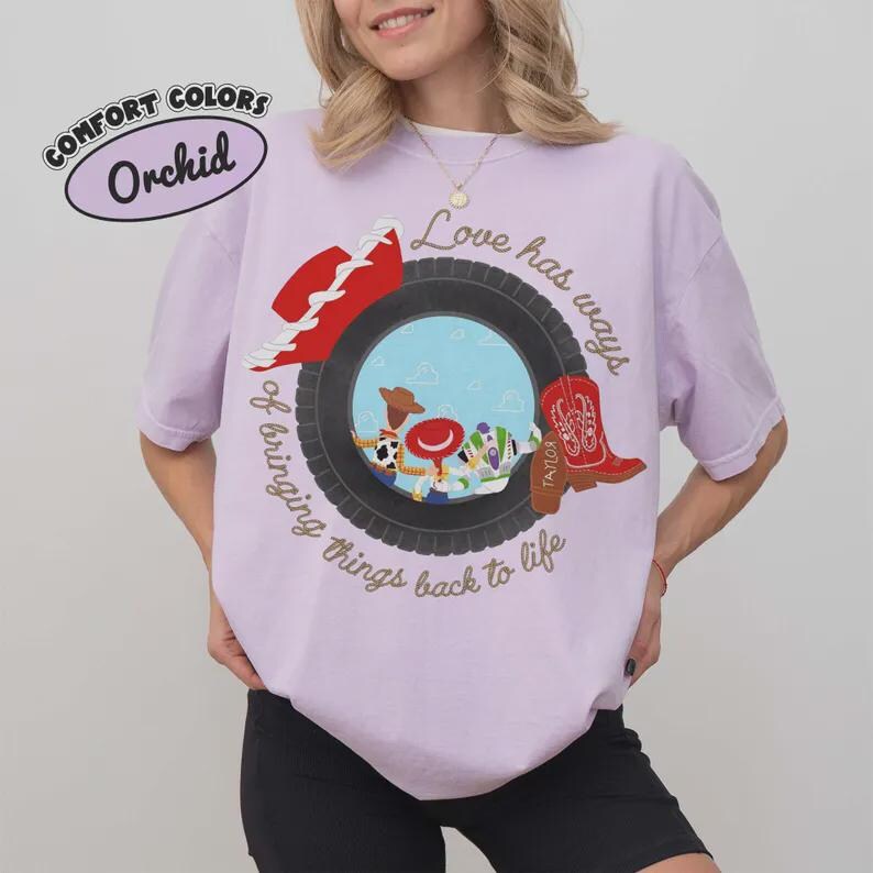 Puede incluir: Una camiseta de manga corta de color morado claro con un gr&aacute;fico de un neum&aacute;tico negro que rodea a personajes de dibujos animados. El texto dice "Love has ways of bringing things back to life" y "Taylor" en una bota vaquera roja.