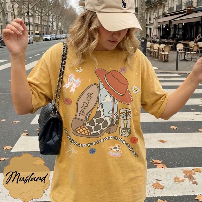 May include: A mustard yellow short-sleeve t-shirt featuring a graphic print of a cowboy boot with a cow print pattern, a red hat, a bow, and blocks with the text "13" and "TS". The design includes the text "TAYLOR" and "I KNEW YOU WERE TROUBLE".
