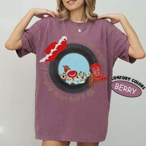 Puede incluir: Una camiseta color baya con un gr&aacute;fico de un neum&aacute;tico negro que enmarca una escena con personajes de dibujos animados. Un sombrero de vaquero rojo y una bota de vaquero roja est&aacute;n sobre el neum&aacute;tico. El texto dice, Love has ways of bringing things back to life.