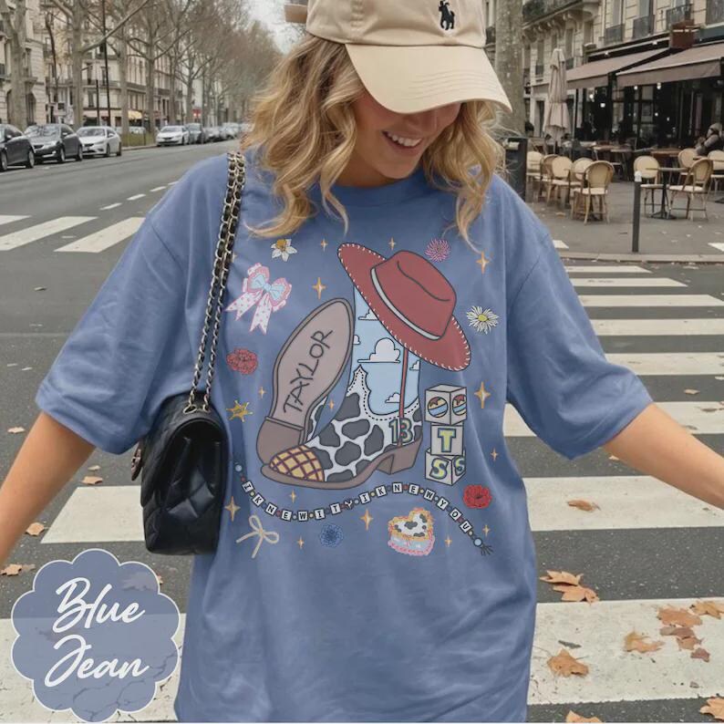 May include: A blue short-sleeve t-shirt featuring a graphic print of a cowboy boot with a cow print toe, a red hat, the name Taylor, the number 13, and stacked blocks with the letters T, S, and S. The design includes small stars, flowers, and bows.