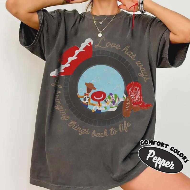 Puede incluir: Una camiseta gris carb&oacute;n de manga corta con un gr&aacute;fico circular que muestra personajes de dibujos animados, una bota de vaquero roja y el texto "Love has ways of bringing things back to life." Camiseta de cuello redondo con efecto lavado.