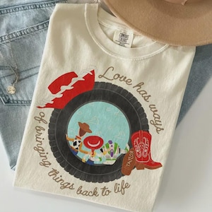 Puede incluir: Una camiseta de color crema con un gr&aacute;fico de un neum&aacute;tico negro que rodea un c&iacute;rculo azul claro con personajes de dibujos animados. El texto dice Love has ways of bringing things back to life. Tambi&eacute;n se muestran un sombrero y una bota vaquera rojos.
