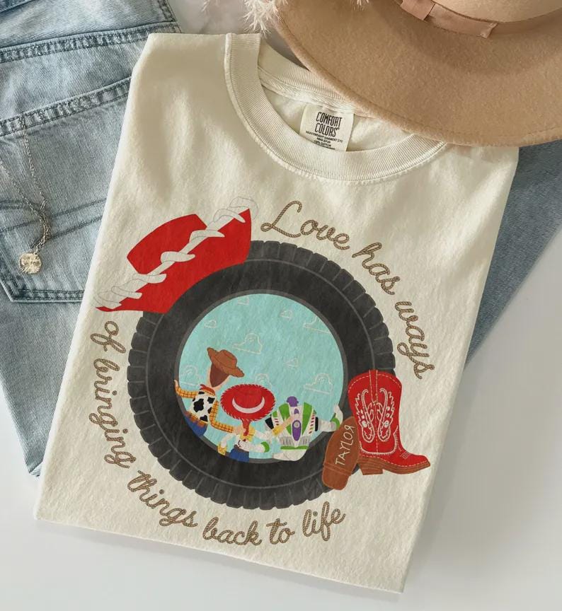 Puede incluir: Una camiseta de color crema con un gr&aacute;fico de un neum&aacute;tico negro que rodea un c&iacute;rculo azul claro con personajes de dibujos animados. El texto dice Love has ways of bringing things back to life. Tambi&eacute;n se muestran un sombrero y una bota vaquera rojos.