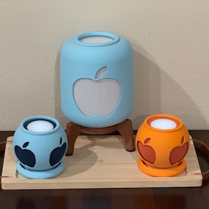 Puede incluir: Un soporte cilíndrico para altavoz de color azul claro con un recorte de manzana, flanqueado por dos soportes esféricos más pequeños en azul claro y naranja, cada uno con recortes de manzana, sobre una bandeja de madera rectangular.