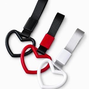 Op de afbeelding: Drie hartvormige sleutelhangers in zwart, rood en wit. Elke sleutelhanger heeft een bijpassende gekleurde band en een zwarte, rode of witte sluiting. De sleutelhangers zijn gerangschikt op een witte achtergrond.