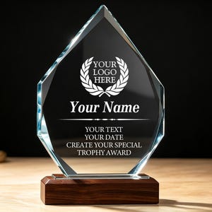 Op de afbeelding: Een ruitvormige trofee van helder glas met een lauwerkransafbeelding en de tekst: YOUR LOGO HERE, Your Name, YOUR TEXT, YOUR DATE, CREATE YOUR SPECIAL TROPHY AWARD. Het glas is bevestigd op een rechthoekige, donkerbruine houten voet.