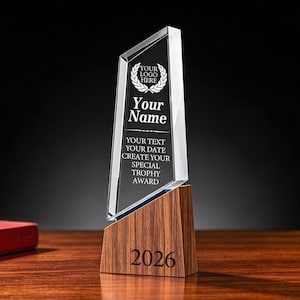 Może przedstawiać: Przezroczyste, kanciaste szklane trofeum osadzone na podstawie z ciemnego drewna. Na szkle wygrawerowano logo wieńca laurowego, napis "Your Name" oraz "YOUR TEXT YOUR DATE CREATE YOUR SPECIAL TROPHY AWARD". Na drewnianej podstawie widnieje rok "2026".